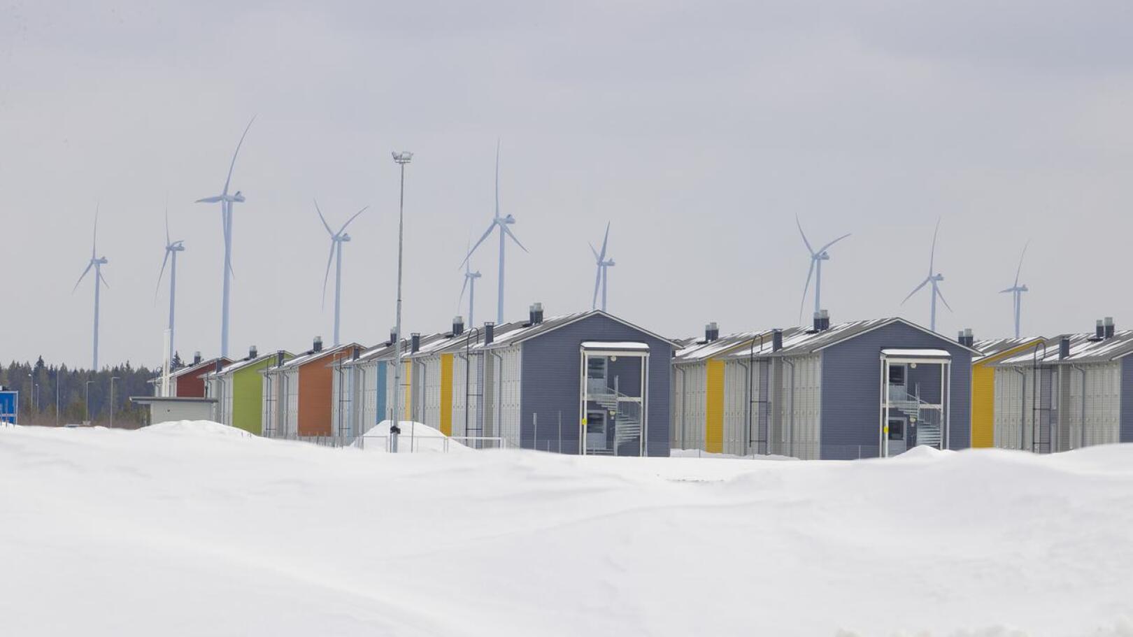 Työntekijät asuvat työmaa-alueen lähellä kerrostaloissa. Taustalla on Pyhäjoella sijaitsevia tuulivoimaloita.