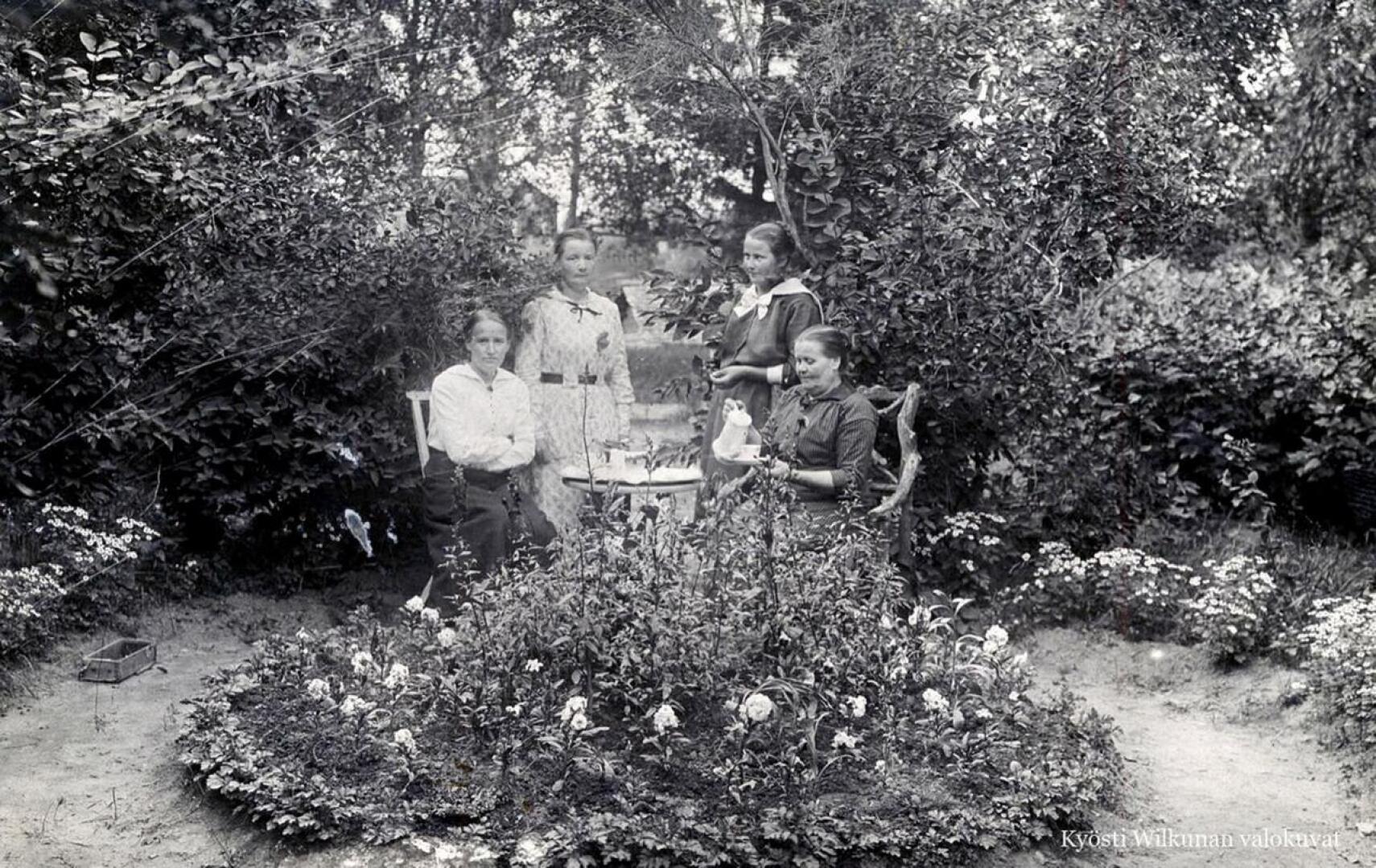 Maikki Kärenlampi istuu kuvassa oikealla taidokkaasti juurakosta valmistetussa tuolissa. Kuva on mahdollisesti otettu hänen puutarhassaan Koivulassa ystävien tai sukulaisten kanssa.  