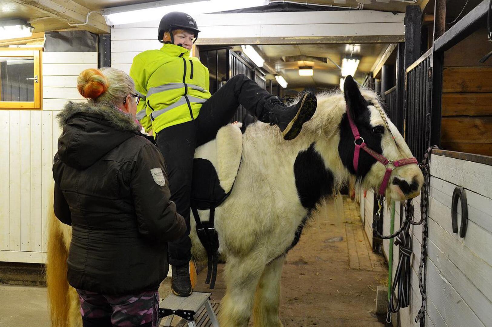 Pia Isoherranen (vas.) ja Heli Somero näyttävät, kuinka asiakas avustetaan hevosen selkään.
